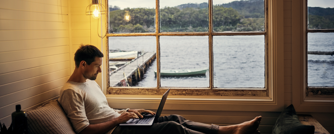 Mann mit Laptop in einem Ferienhaus vor Seekulisse mit Boot.